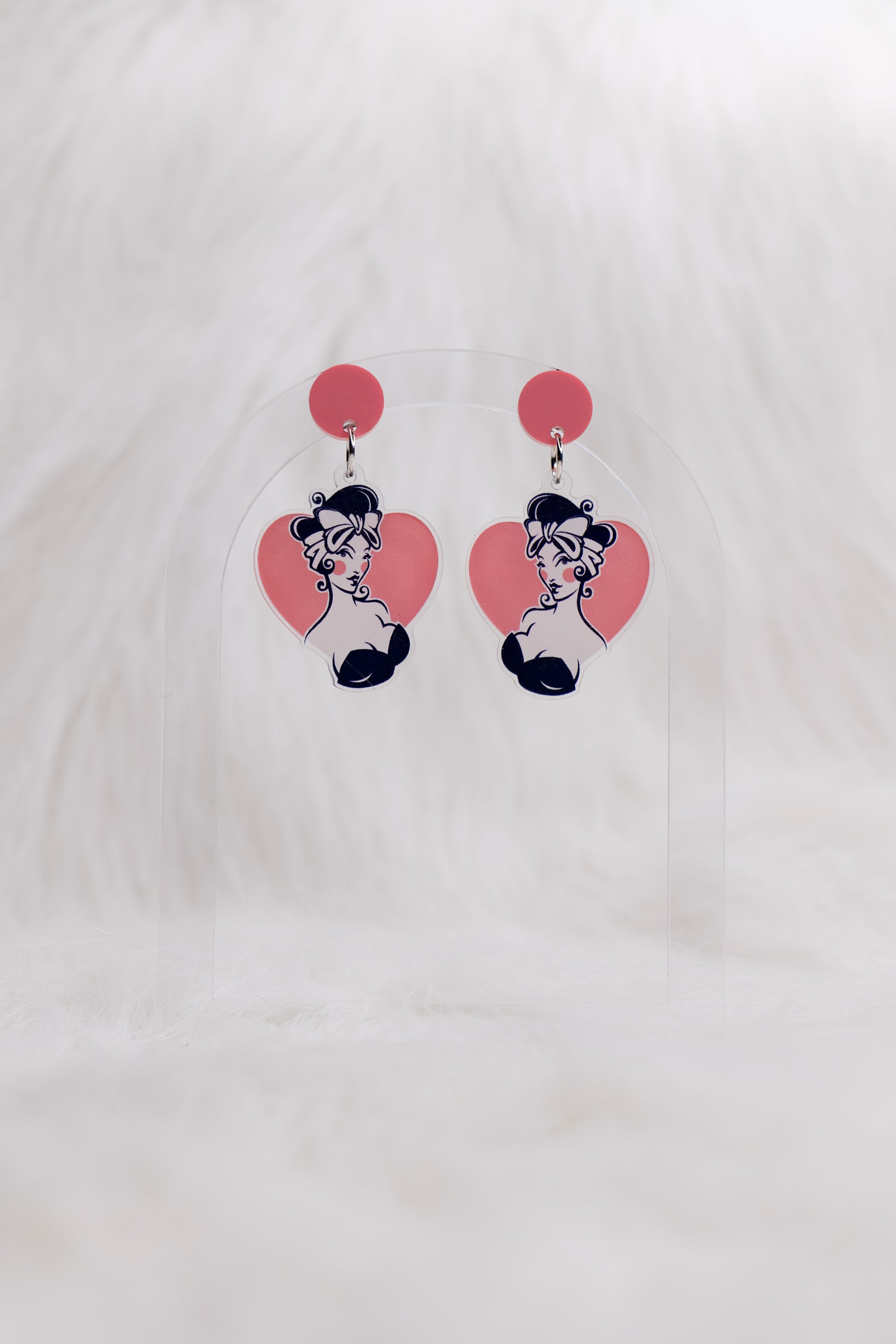 Pair of acrylic earrings with a pink circular stud and heart-shaped drops featuring a retro pin-up styled illustration of a girl with dark hair and bow; lightweight with stainless steel posts and large backs for accessibility, displayed against a white backgound 