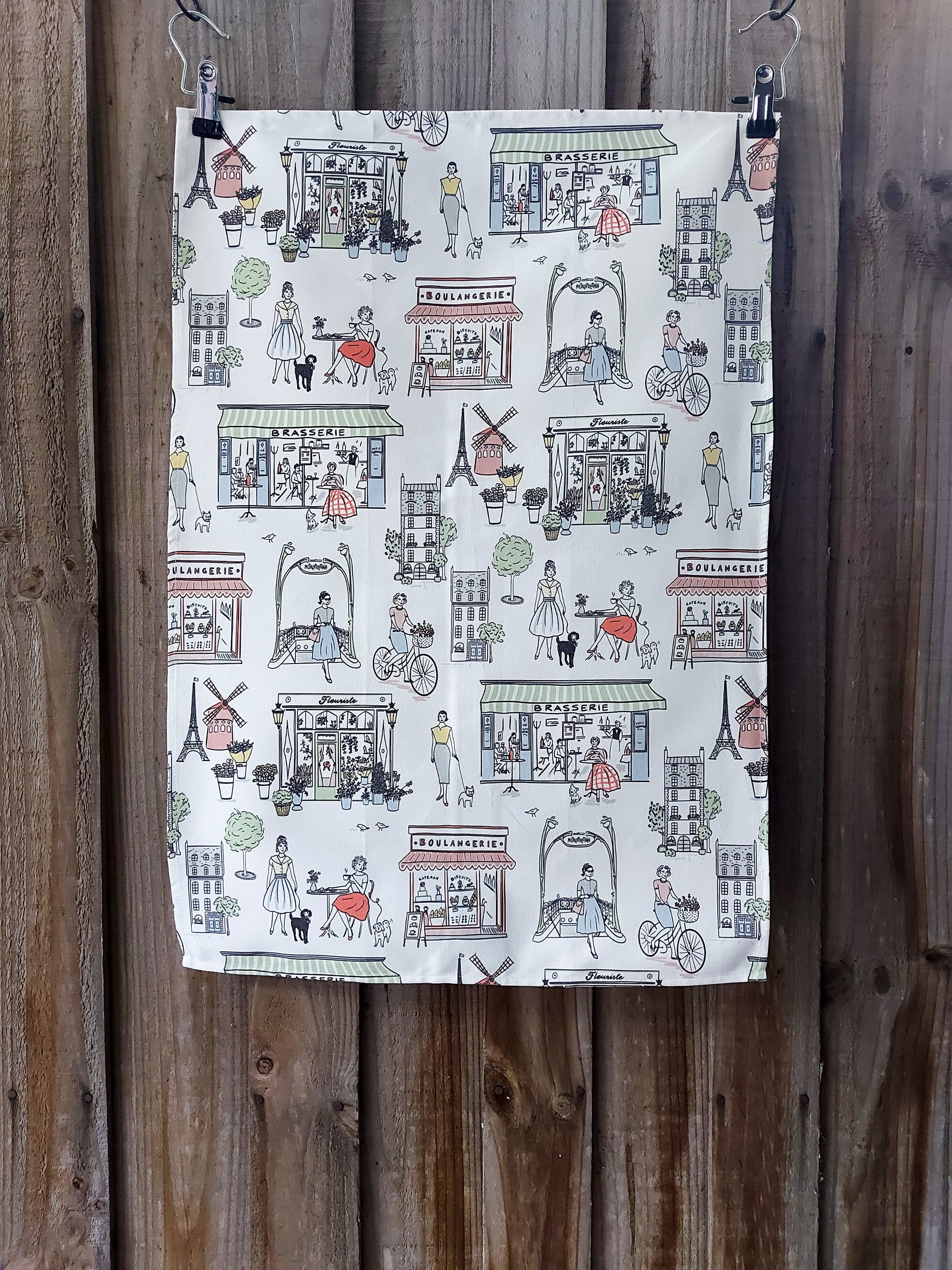100% cotton tea towel featuring retro Parisian illustrations in pastel colours on a white background; displayed against a wooden fence.