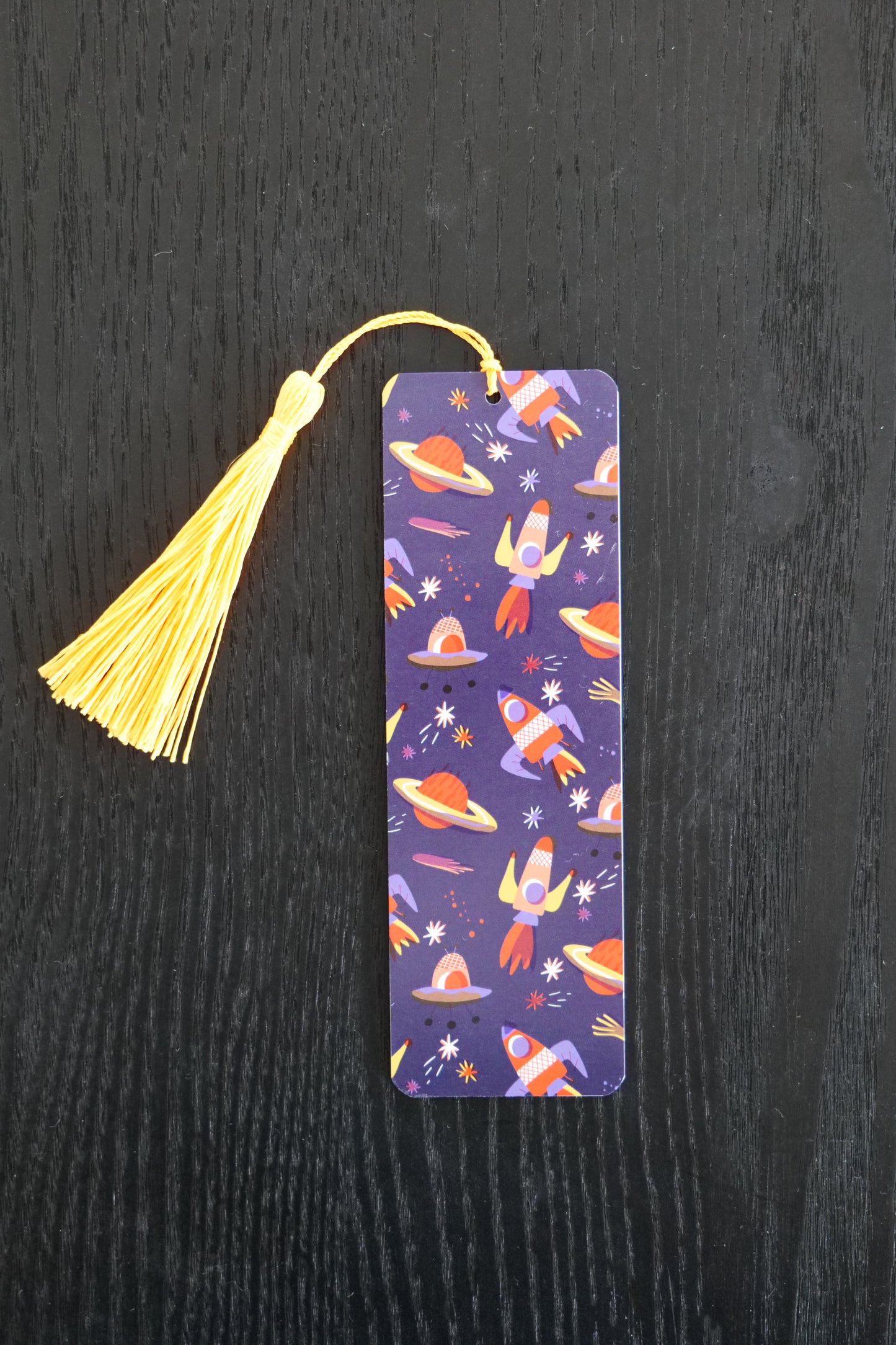 Bookmark showing red and yellow rockets on a dark blue background with a yellow tassel, sitting against a black backdrop