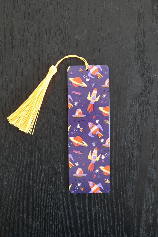 Bookmark showing red and yellow rockets on a dark blue background with a yellow tassel, sitting against a black backdrop
