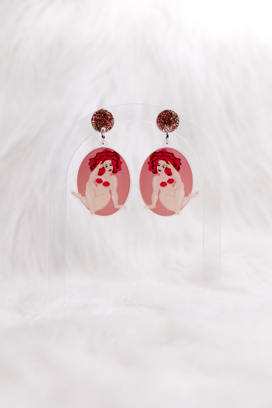 Pair of lightweight acrylic earrings in an oval shape, featuring an illustration of a nude woman with red hair and strategically placed red frosting on a pink background, earrings have stainless steel posts and large accessible back, displayed against a white backdrop