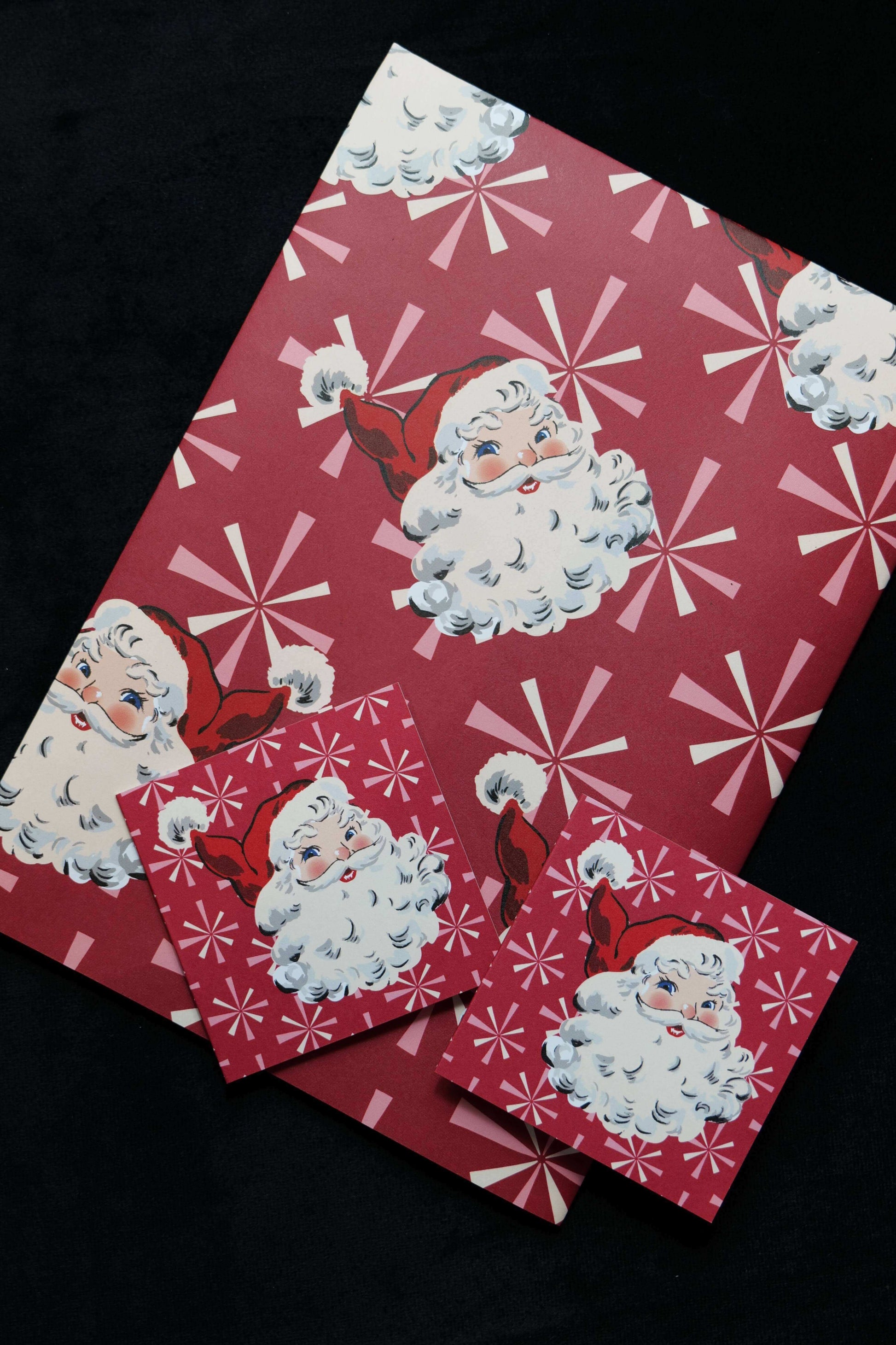 A folded sheet of gift wrap featuring retro style Santa faces on a red background with windmill stars, also showing two matching cards, displayed against a black backdrop