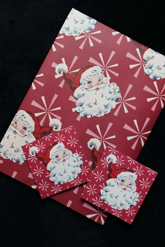 A folded sheet of gift wrap featuring retro style Santa faces on a red background with windmill stars, also showing two matching cards, displayed against a black backdrop