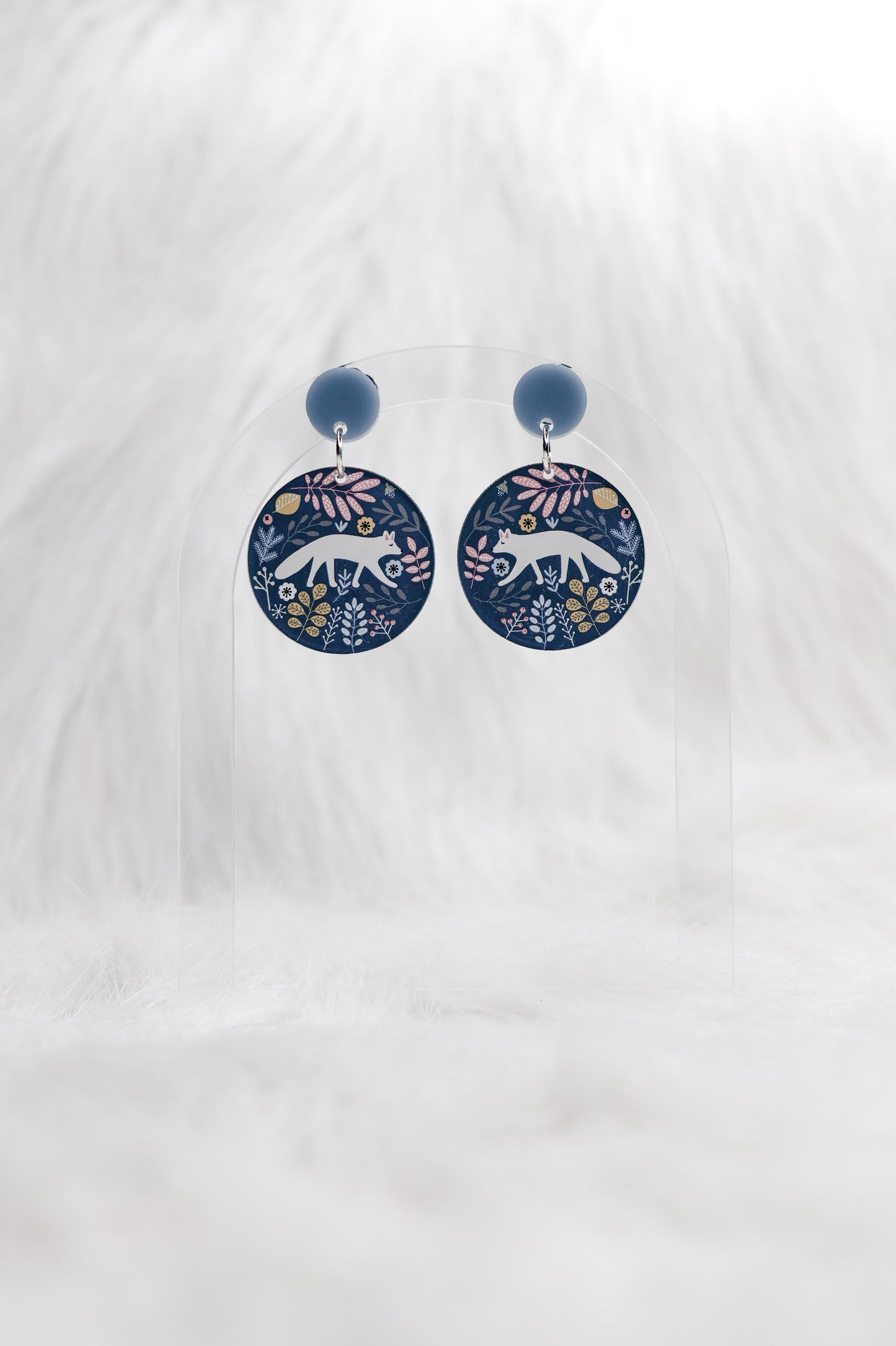 Round earrings featuring a stylised white fox and leaves in blue and beige on a grey background and grey blue stud top, displayed on a clear stand against a white background