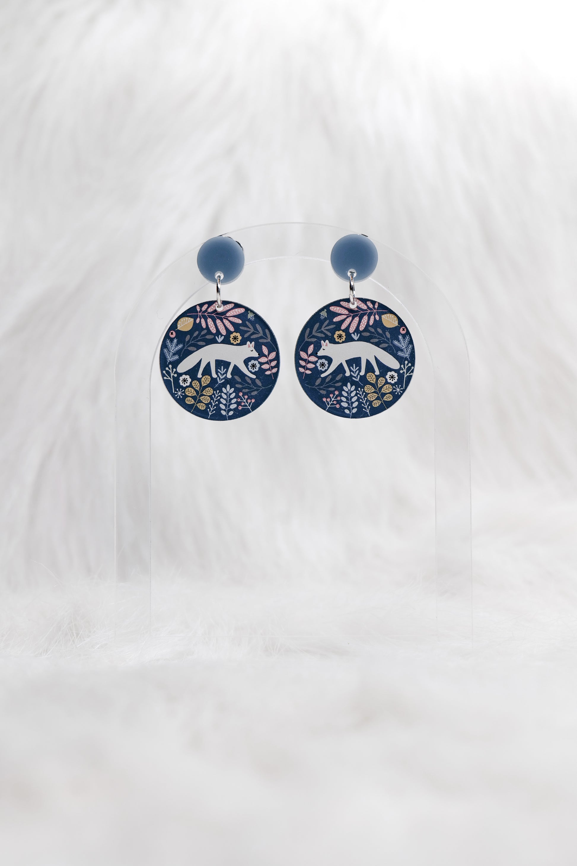 Round earrings featuring a stylised white fox and leaves in blue and beige on a grey background and grey blue stud top, displayed on a clear stand against a white background