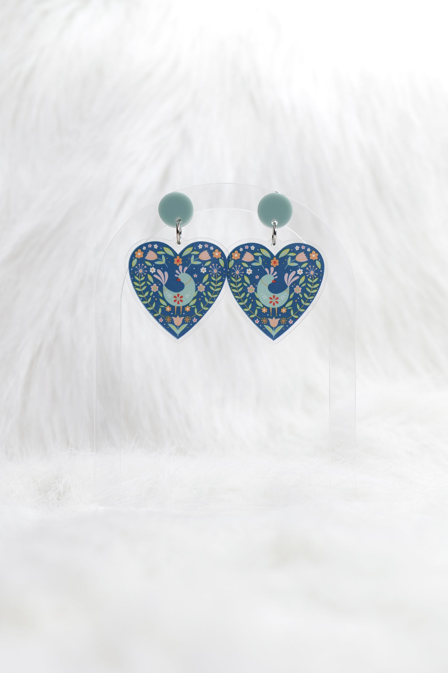 Heart shaped earrings in deep teal featuring a central bird surrounded by Scandinavian-inspired floral designs, hanging under a light teal stud; displayed on a clear stand against a white background.