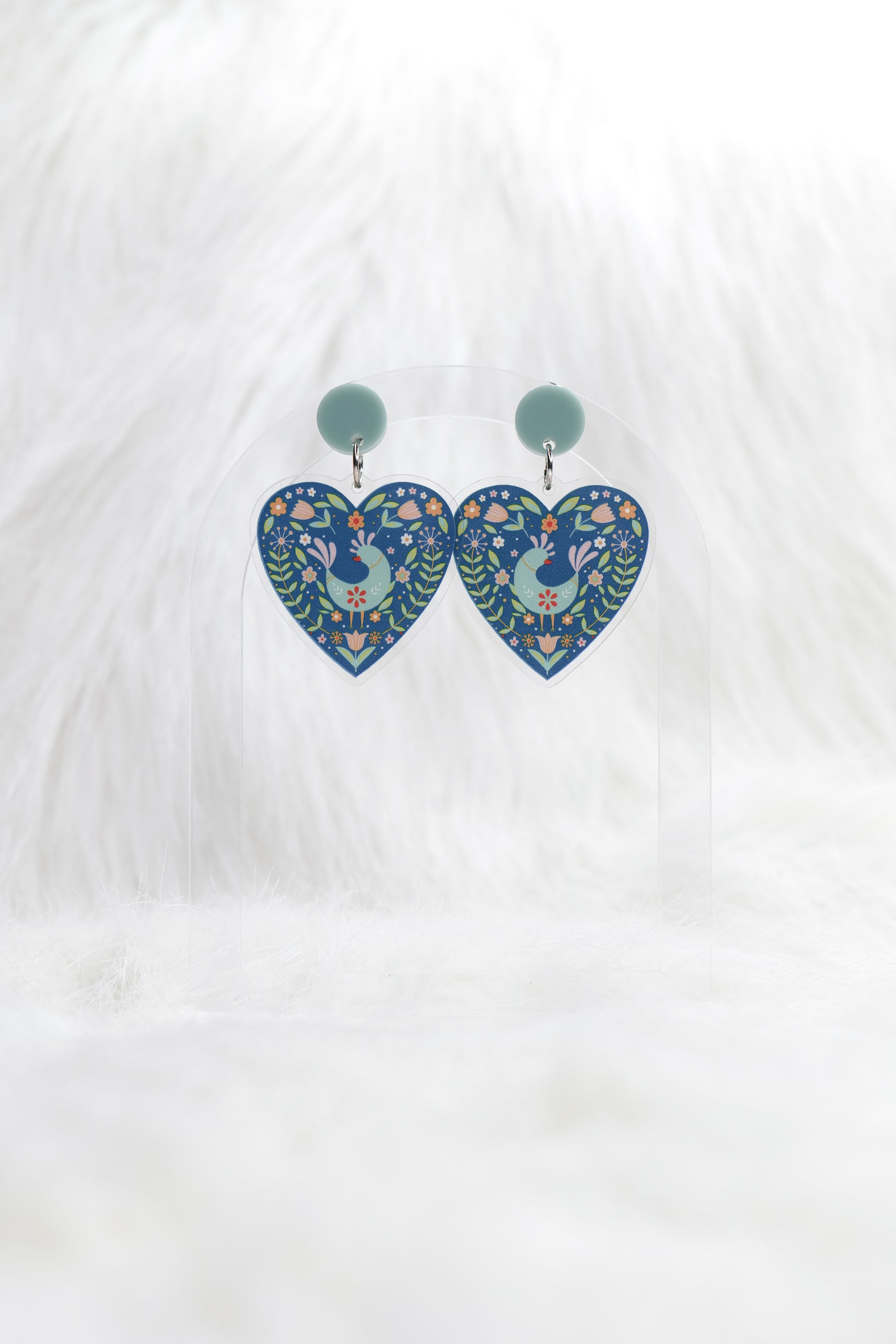 Heart shaped earrings in deep teal featuring a central bird surrounded by Scandinavian-inspired floral designs, hanging under a light teal stud; displayed on a clear stand against a white background.