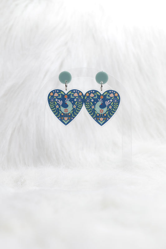 Heart shaped earrings in deep teal featuring a central bird surrounded by Scandinavian-inspired floral designs, hanging under a light teal stud; displayed on a clear stand against a white background.