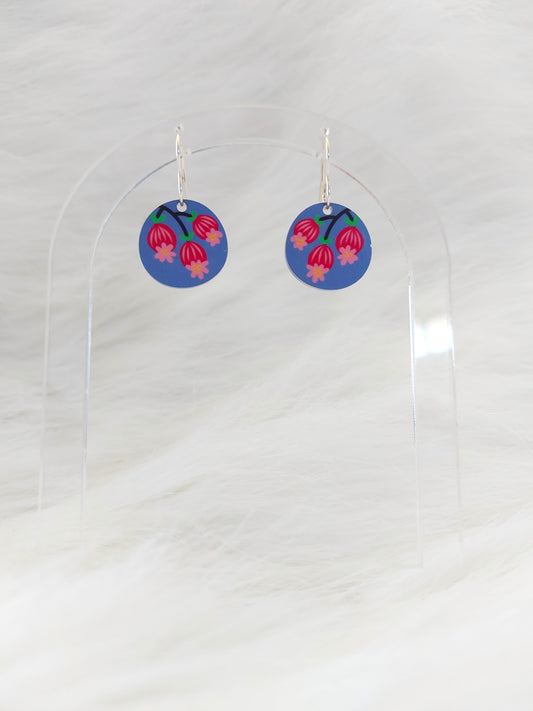 Pair of round earrings featuring three Scandinavian inspired blossoms against a blue-mauve background; printed on lightweight acrylic with stainless steel hooks, displayed against a white background