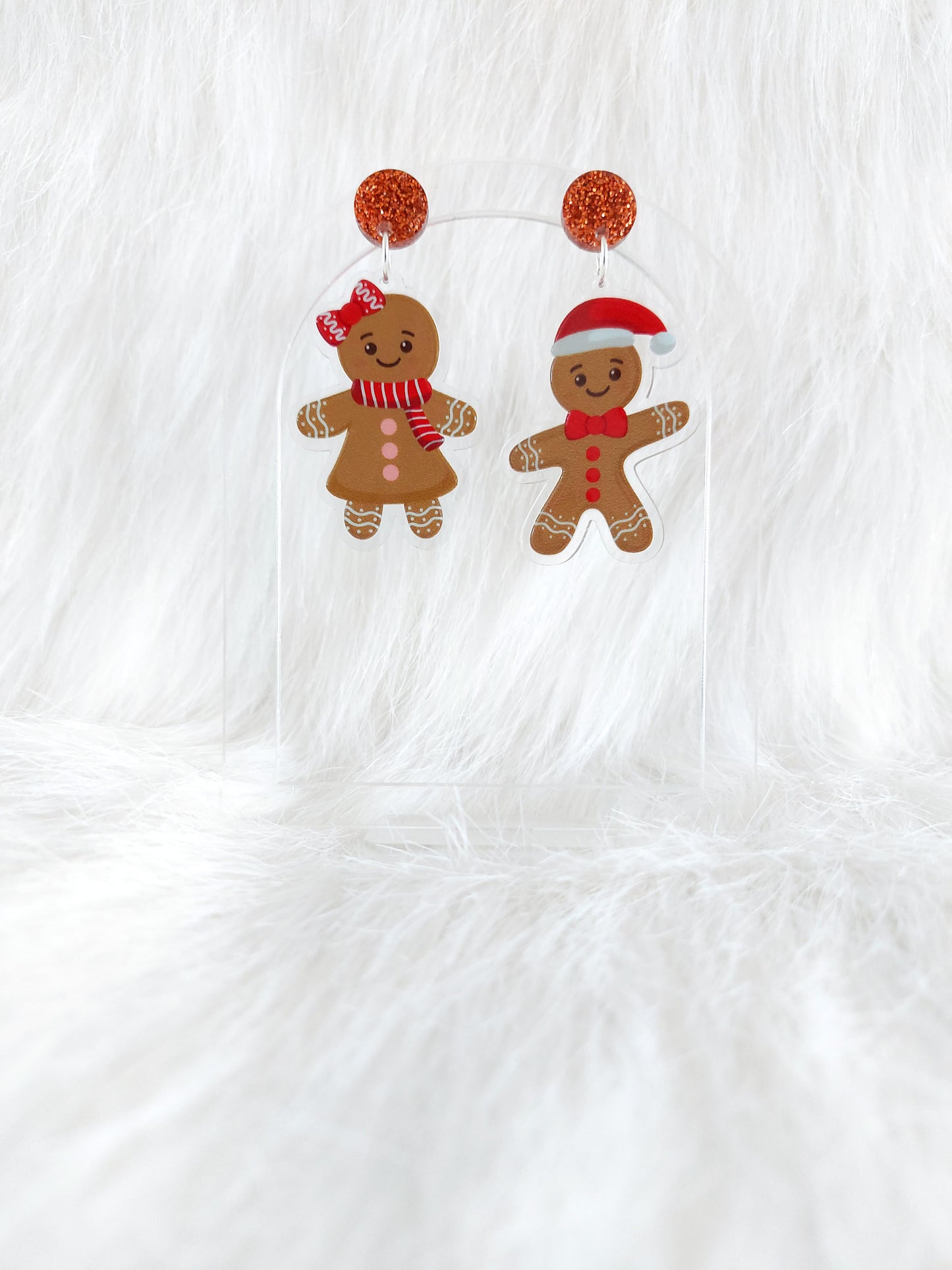 A pair of gingerbread character earrings, one male wearing a Santa hat and one female wearing a red bow and scarf, hanging from a copper glitter top, displayed on a clear stand in front of a white background