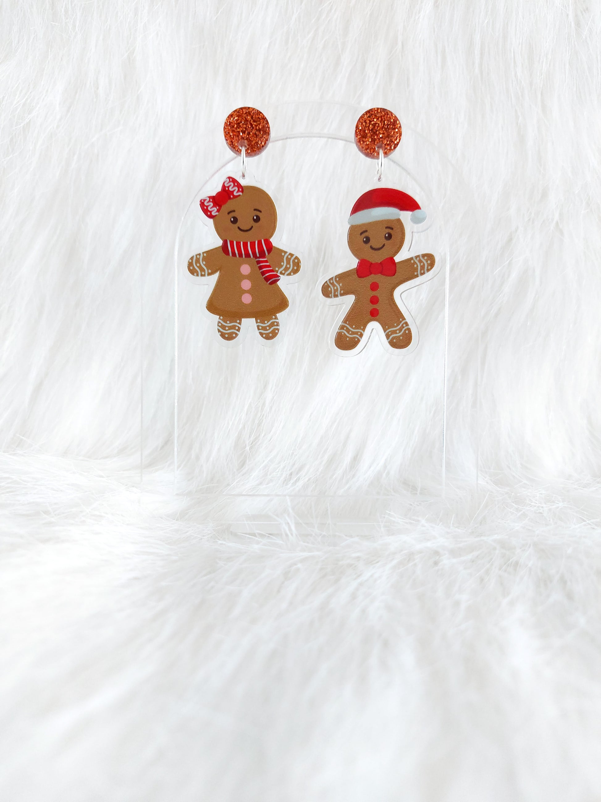 A pair of gingerbread character earrings, one male wearing a Santa hat and one female wearing a red bow and scarf, hanging from a copper glitter top, displayed on a clear stand in front of a white background