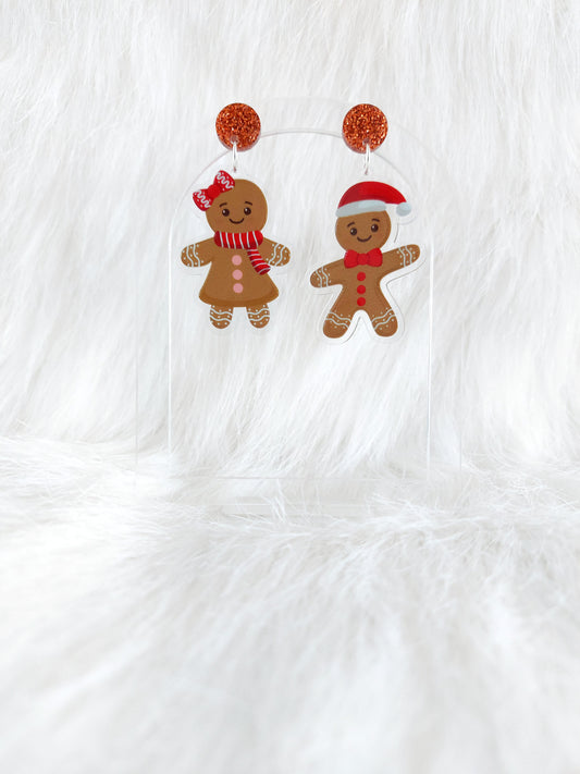 A pair of gingerbread character earrings, one male wearing a Santa hat and one female wearing a red bow and scarf, hanging from a copper glitter top, displayed on a clear stand in front of a white background