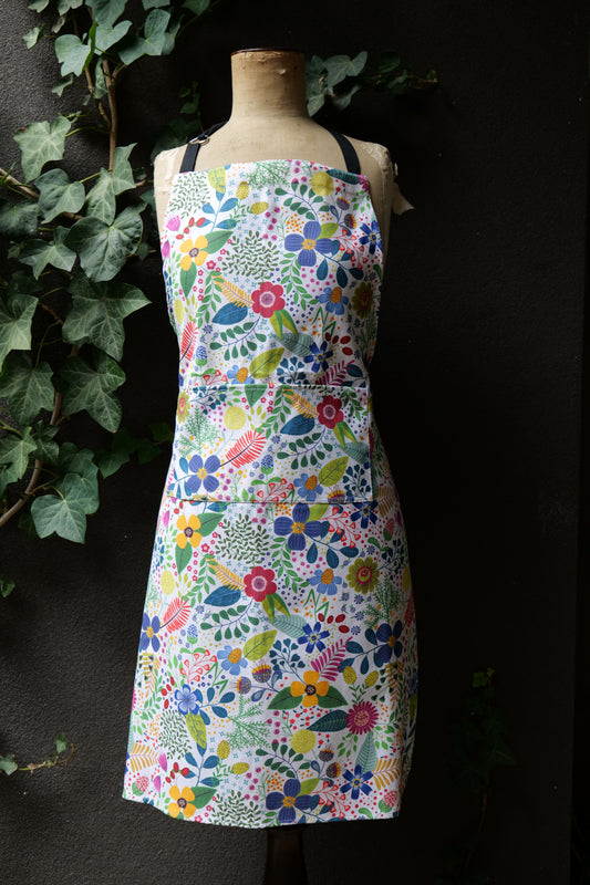 Bright floral Spring Garden apron in pink, yellow, and green tones, featuring a front pocket, adjustable neck ties, and long waist ties. Made from premium heavyweight cotton, Australian made, shown on a mannequin against a dark background.