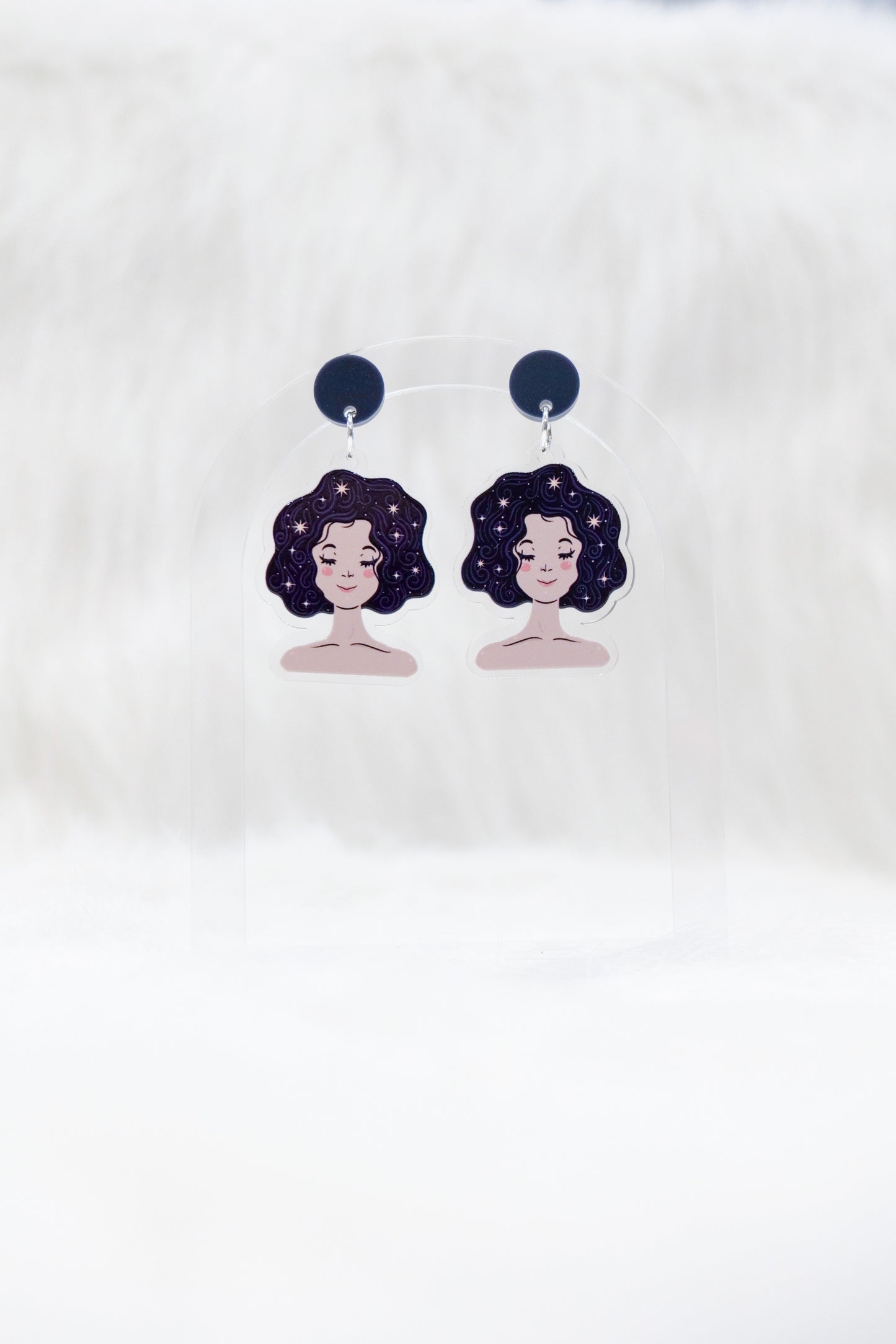 A pair of acrylic earrings featuring a sleeping girl's face with stars in her hair and dark charcoal stud tops, displayed on a clear stand against a white background