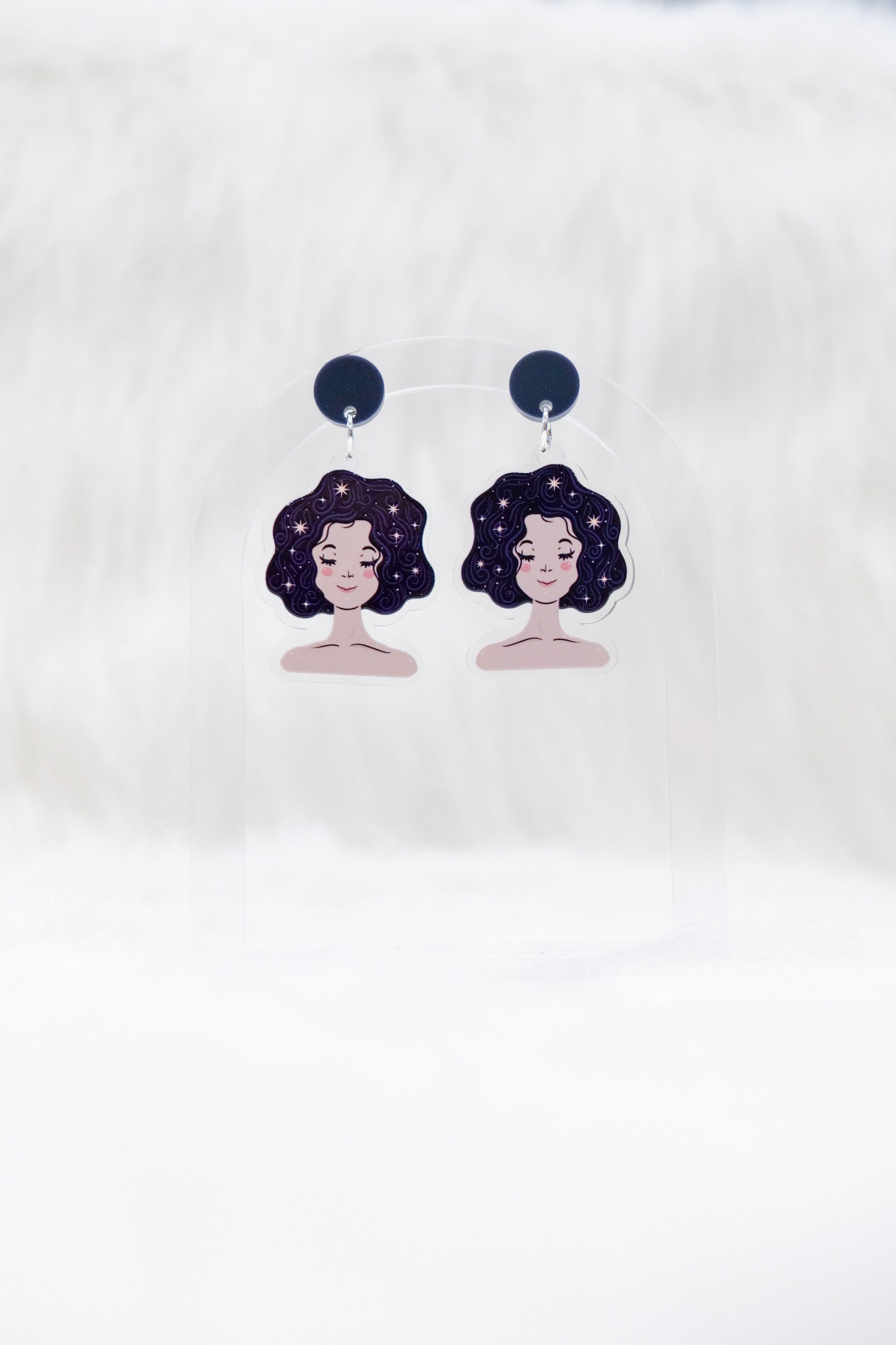 A pair of acrylic earrings featuring a sleeping girl's face with stars in her hair and dark charcoal stud tops, displayed on a clear stand against a white background