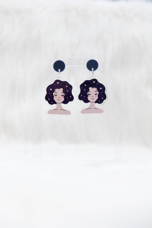 A pair of acrylic earrings featuring a sleeping girl's face with stars in her hair and dark charcoal stud tops, displayed on a clear stand against a white background