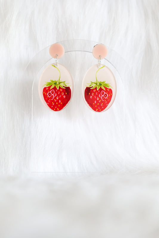 Oval earrings with pale pink studs featuring bold graphic style strawberries with a green top on a pale pink background, displayed on a clear stand against a white backdrop