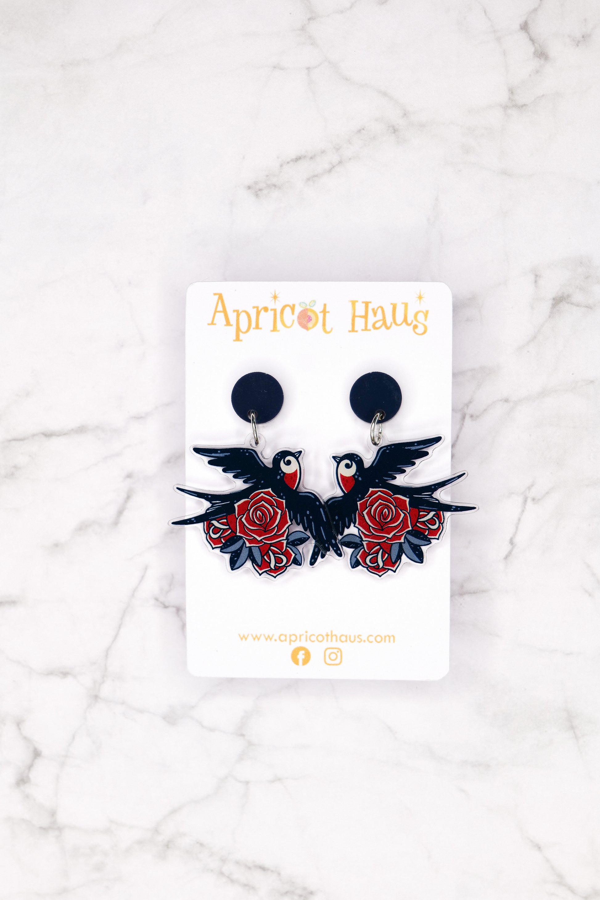 Pair of acrylic earrings featuring dark navy swallows with red chests surrounded by red roses in old tattoo style; lightweight with stainless steel posts for hypoallergenic wear; displayed on an Apricot Haus backing card.