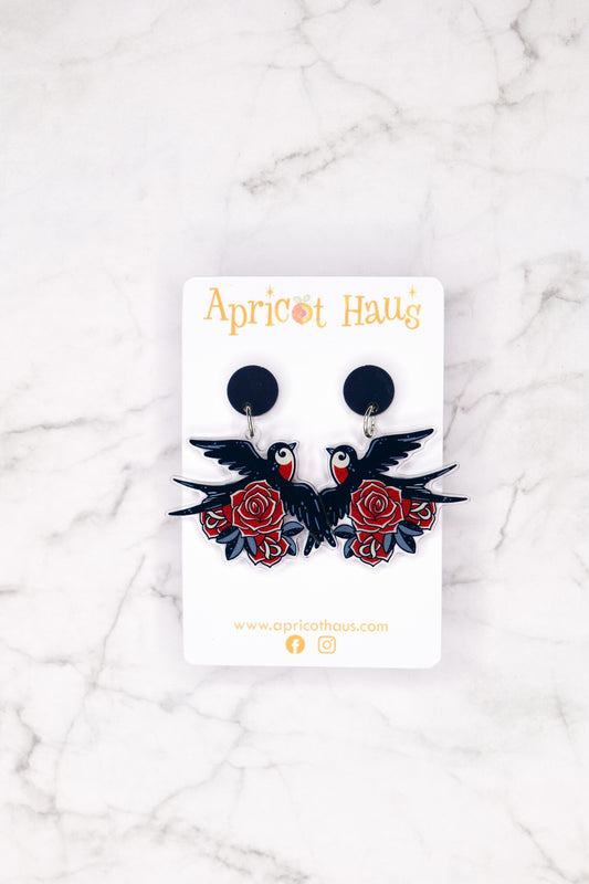 Pair of acrylic earrings featuring dark navy swallows with red chests surrounded by red roses in old tattoo style; lightweight with stainless steel posts for hypoallergenic wear; displayed on an Apricot Haus backing card.