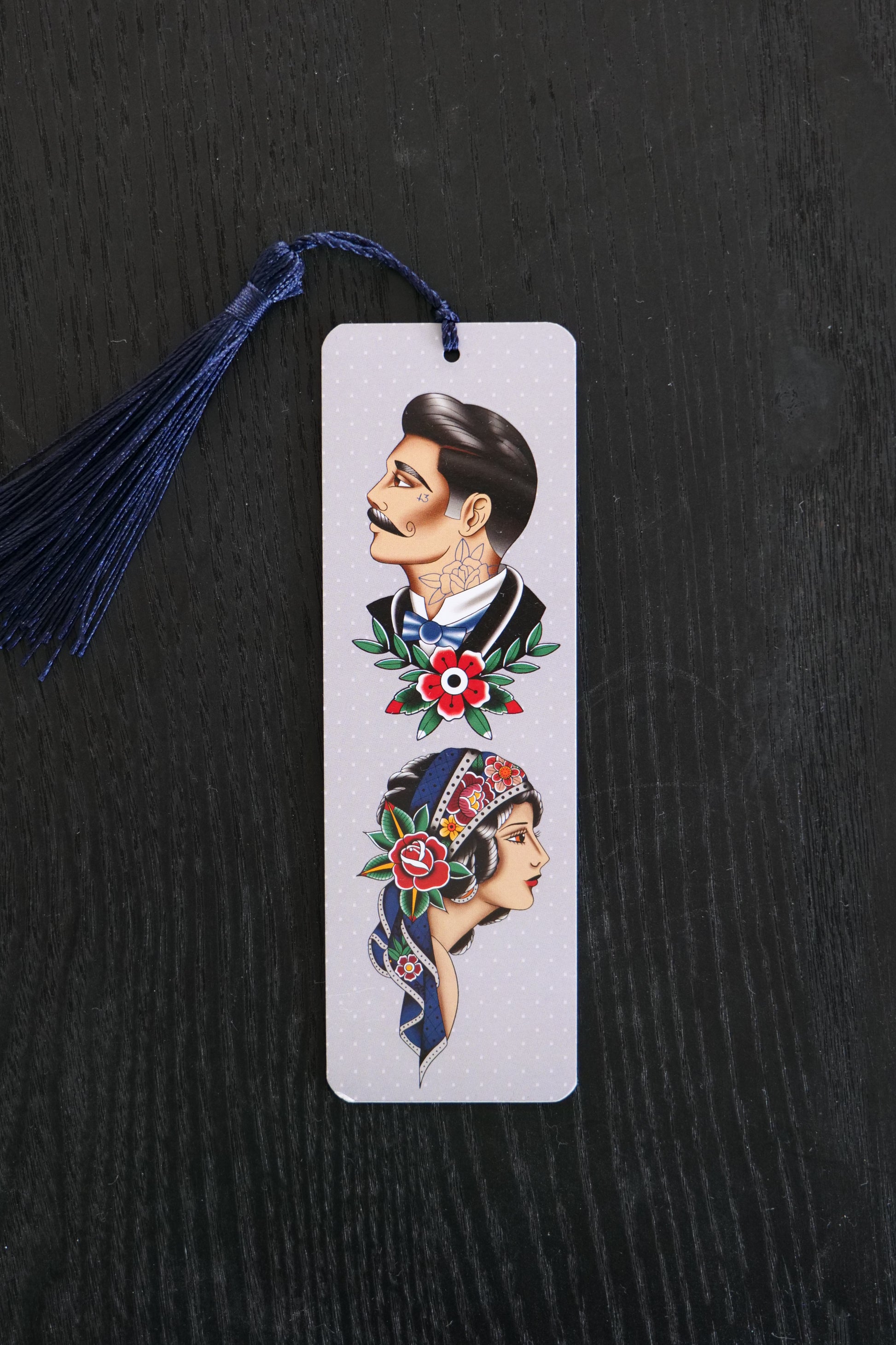 Bookmark with traditional tattoo style male and female faces, against a pale grey background with a navy tassel; sitting against a black backdrop