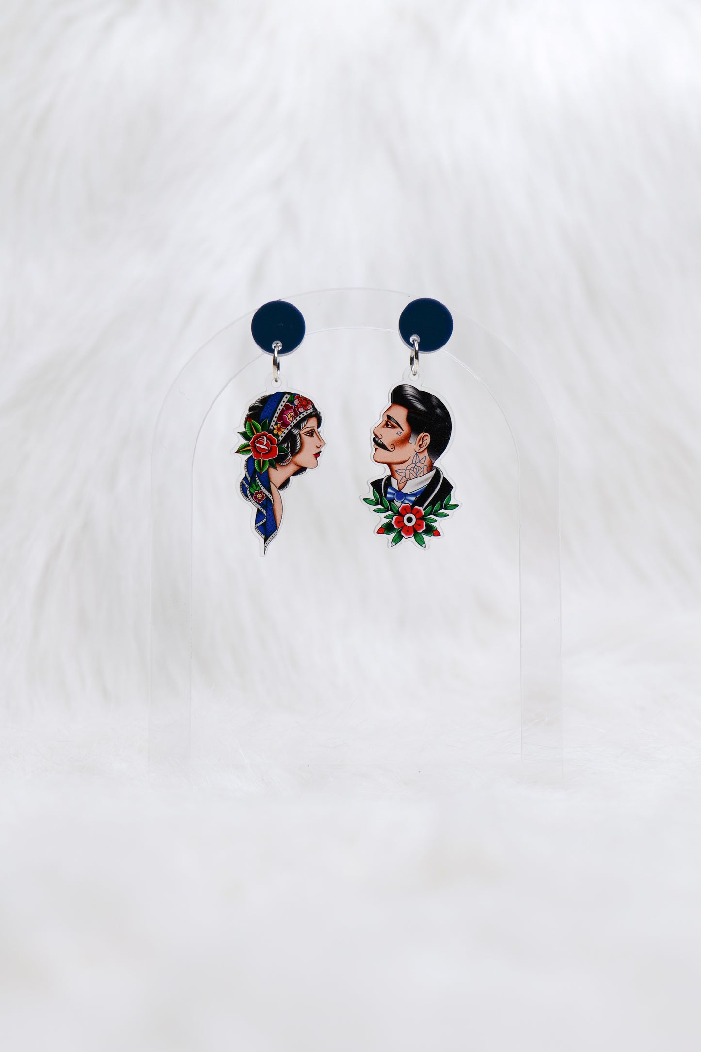 Pair of acrylic earrings featuring hand-drawn tattoo-style portraits: one of a woman with a headscarf and rose, the other of a man with a moustache and traditional tattoo motifs; attached to dark blue circular studs; lightweight with stainless steel posts for comfortable, hypoallergenic wear.