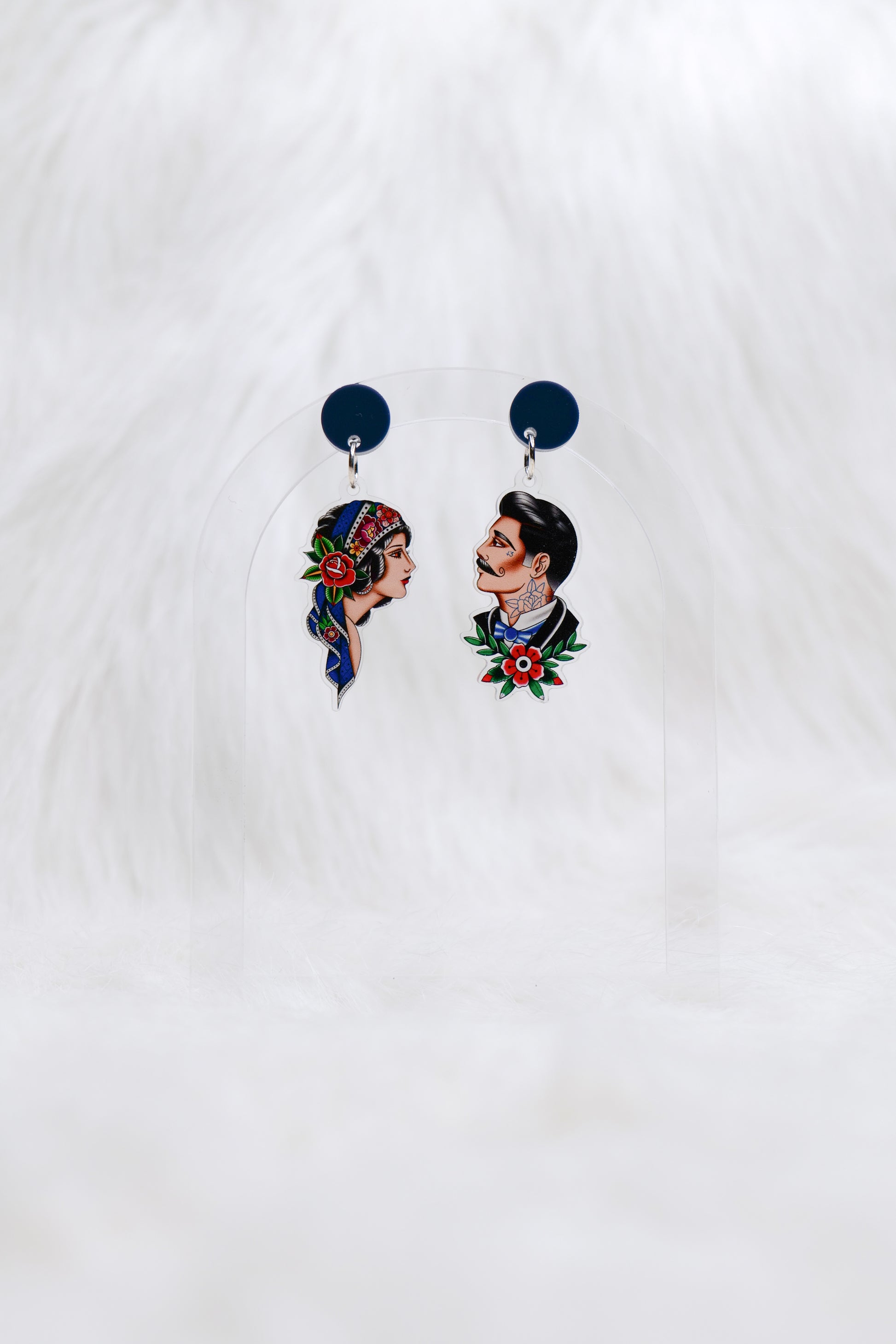 Pair of acrylic earrings featuring hand-drawn tattoo-style portraits: one of a woman with a headscarf and rose, the other of a man with a moustache and traditional tattoo motifs; attached to dark blue circular studs; lightweight with stainless steel posts for comfortable, hypoallergenic wear.