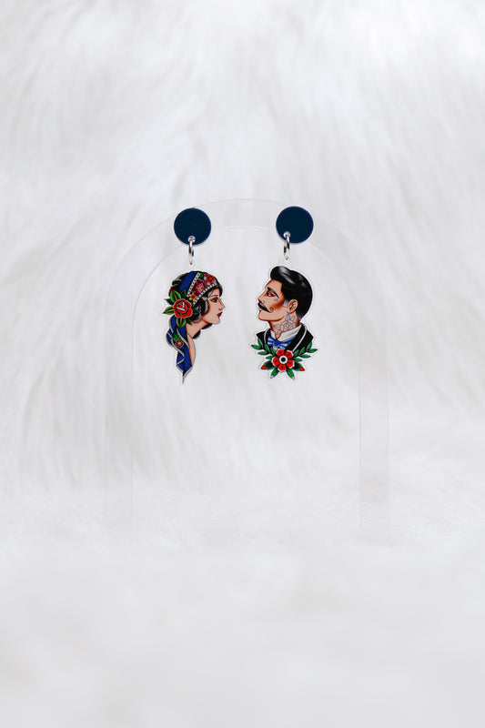 Pair of acrylic earrings featuring hand-drawn tattoo-style portraits: one of a woman with a headscarf and rose, the other of a man with a moustache and traditional tattoo motifs; attached to dark blue circular studs; lightweight with stainless steel posts for comfortable, hypoallergenic wear.
