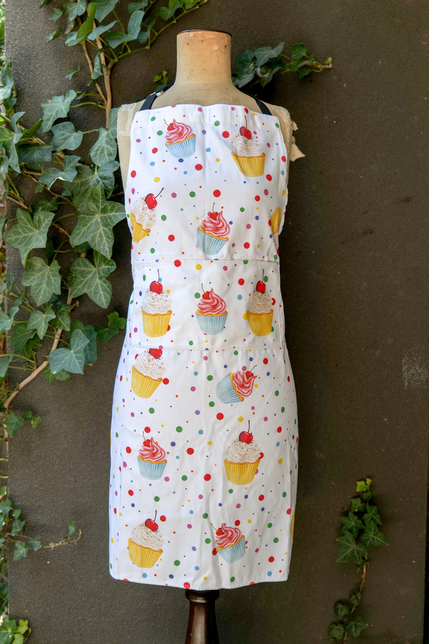 White apron featuring colourful cupcake illustrations with frosting swirls, cherries, and multicoloured polka dots; includes adjustable neck strap, extended waist ties and front pocket; generous fit, handmade in Australia from cotton canvas and machine washable; matching tea towel available.