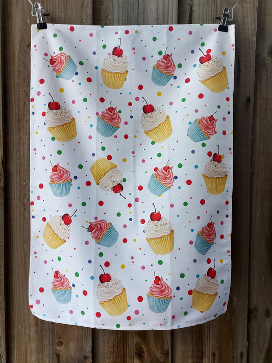 100% cotton tea towel featuring colourful illustrated cupcakes and multicoloured polka dots on a white background; displayed against a wooden fence