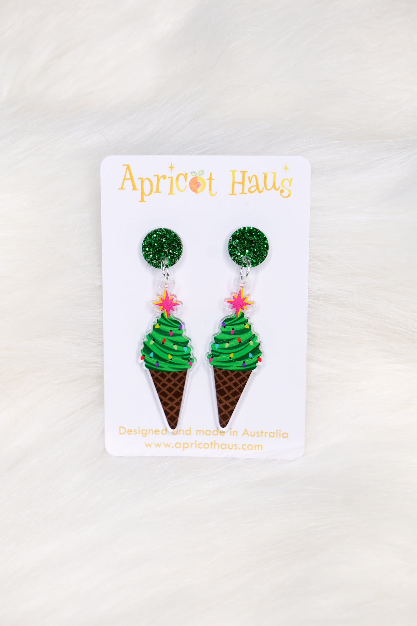 Pair of earrings in the shape of an ice cream, but made of a Christmas tree as the ice cream with a cone base and a green glitter stud top; displayed on an Apricot Haus earring card on a white background