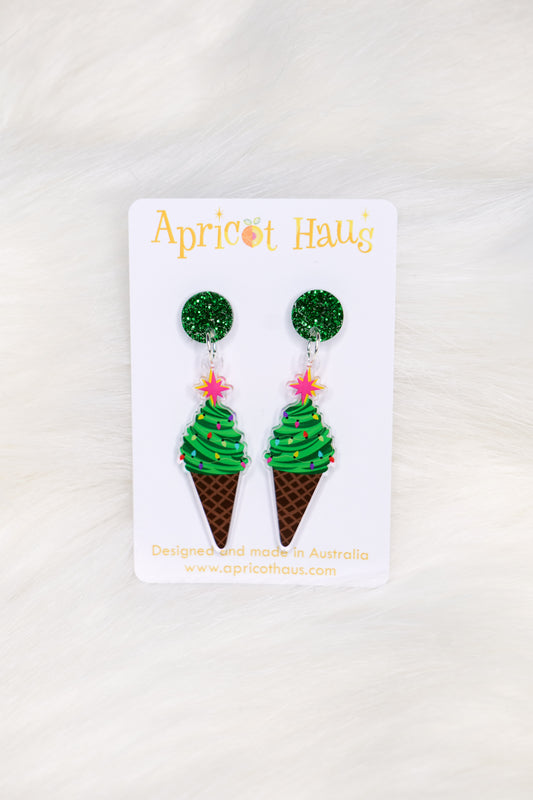 Pair of earrings in the shape of an ice cream, but made of a Christmas tree as the ice cream with a cone base and a green glitter stud top; displayed on an Apricot Haus earring card on a white background