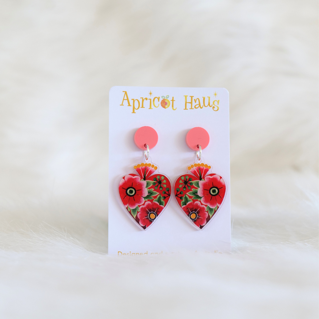 Heart shaped earrings with pink stud tops featuring a red and green watercolour floral design, displayed on an Apricot Haus earring card against a white background