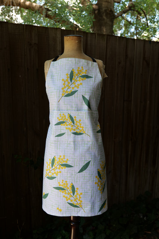 Wattle apron featuring sprigs of wattle blossom and green leaves on pale green check background, modelled on a mannequin in front of a dark background; apron has an adjustable neck and long waist ties, designed and made in Australia, machine washable.