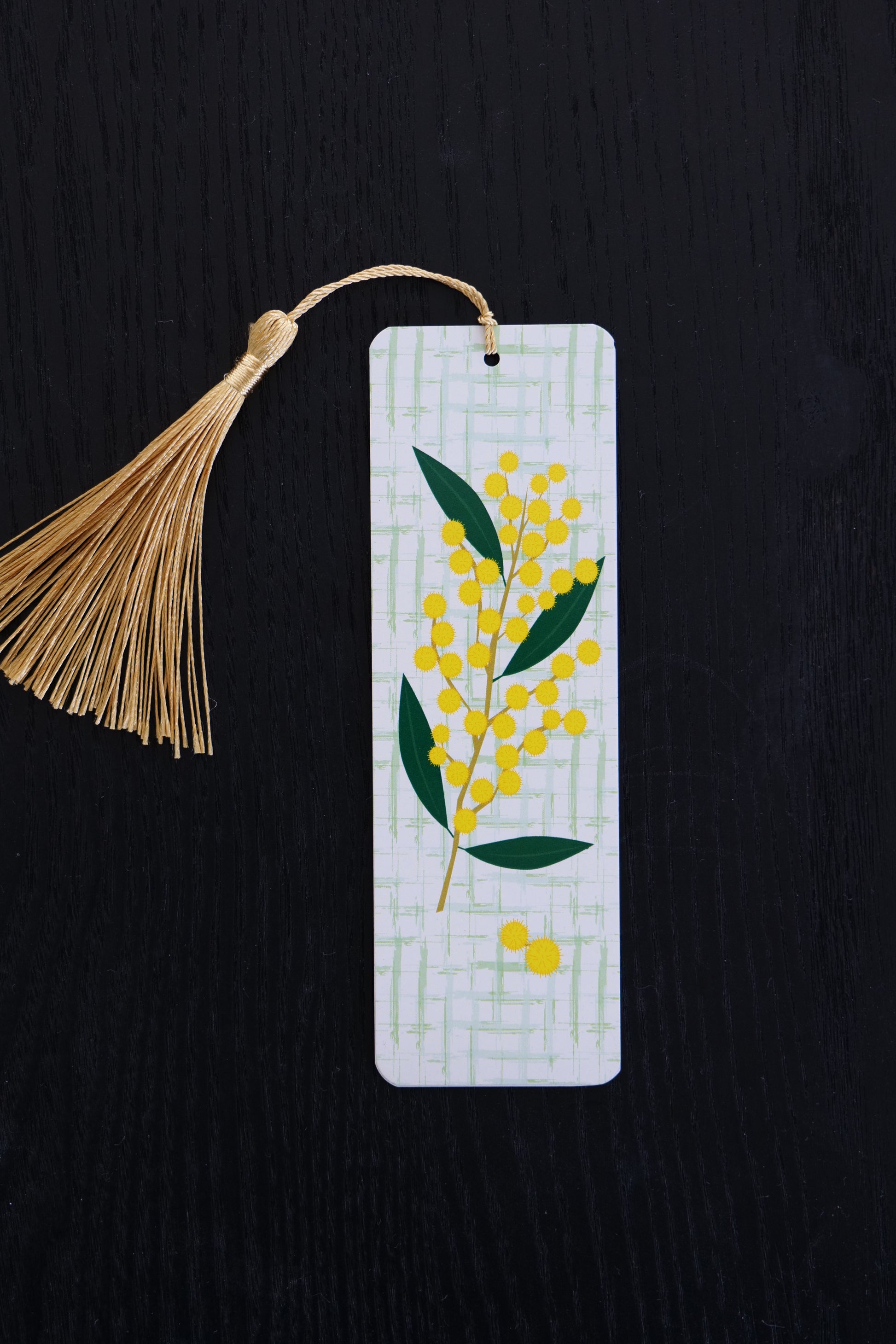 Bookmark featuring a spring of wattle with a green and white lattice background and a gold tassel, sitting against a black backdrop
