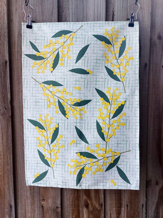 100% cotton tea towel featuring illustrated golden wattle blossoms and green foliage on a light green checked background; displayed against a wooden fence