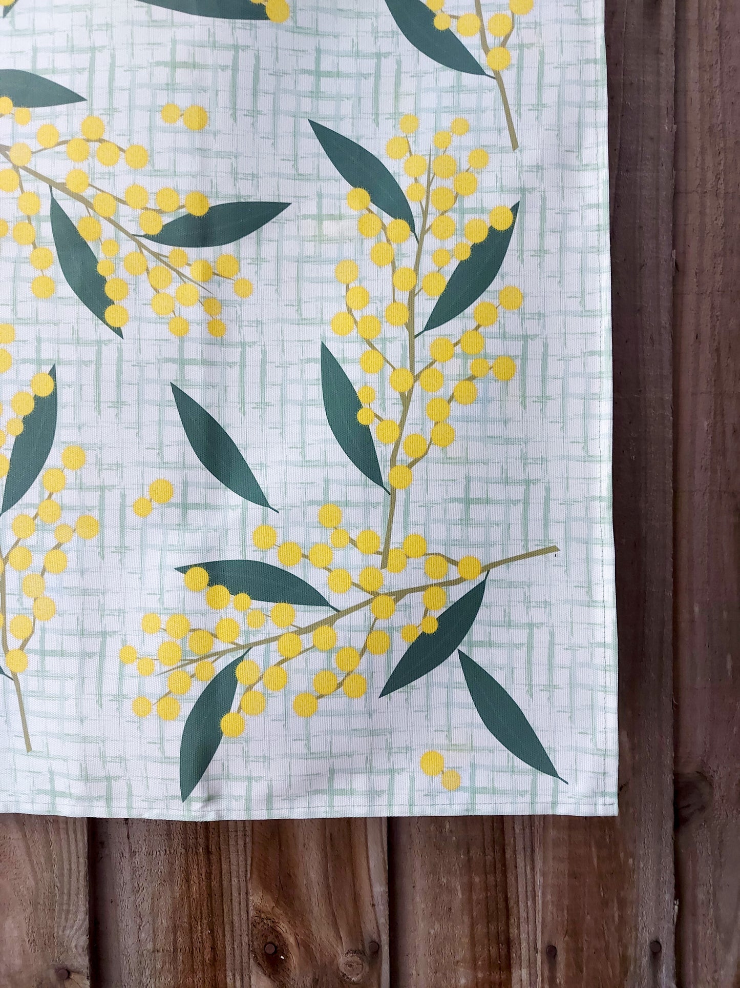 Close up photo of a corner of the wattle tea towel, showing the wattle illustrations on a pale green check background