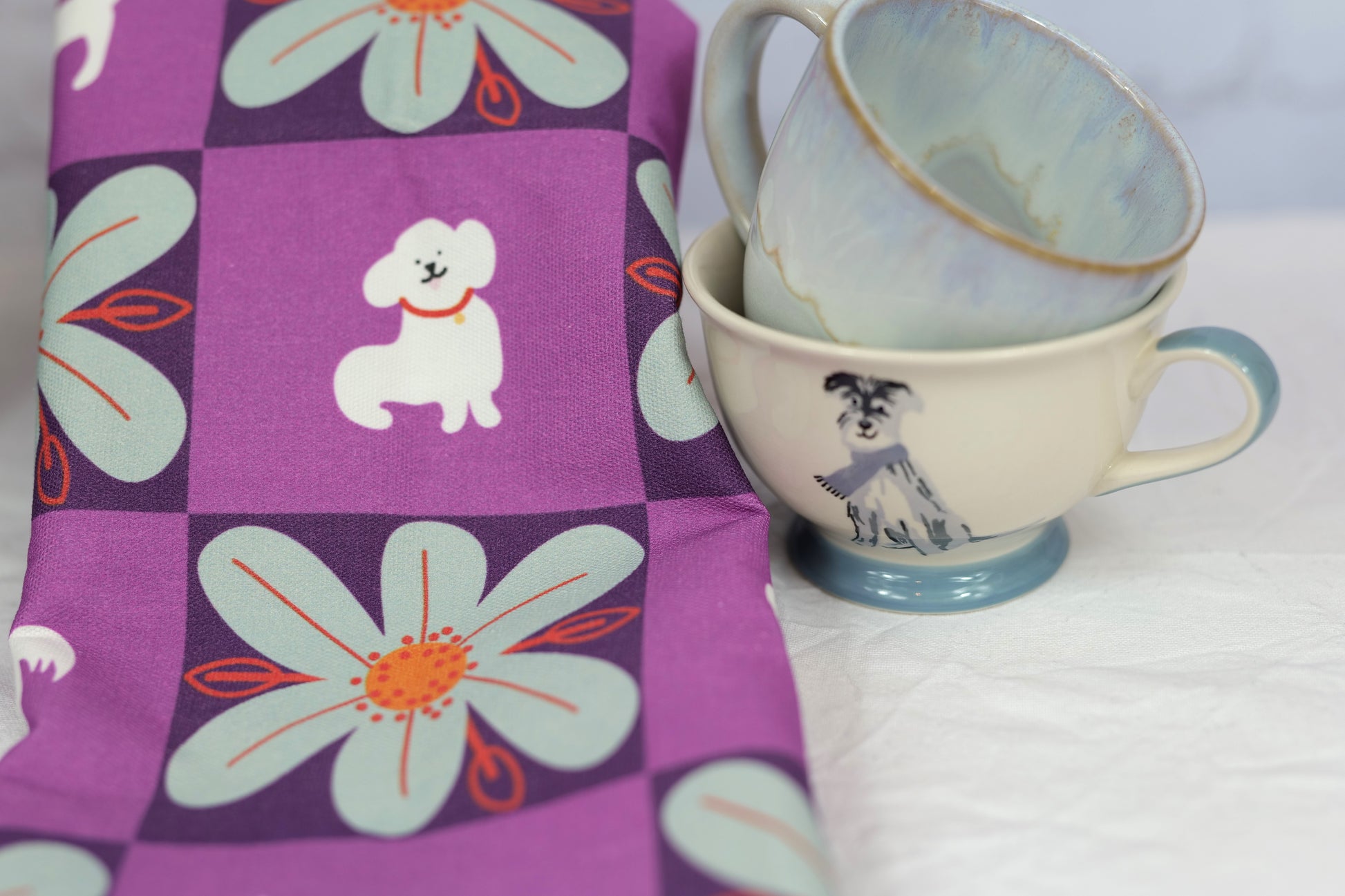 close up photo of the My Hero Winky tea towel, showing the white dog and pale blue flowers, next to two coffee cups